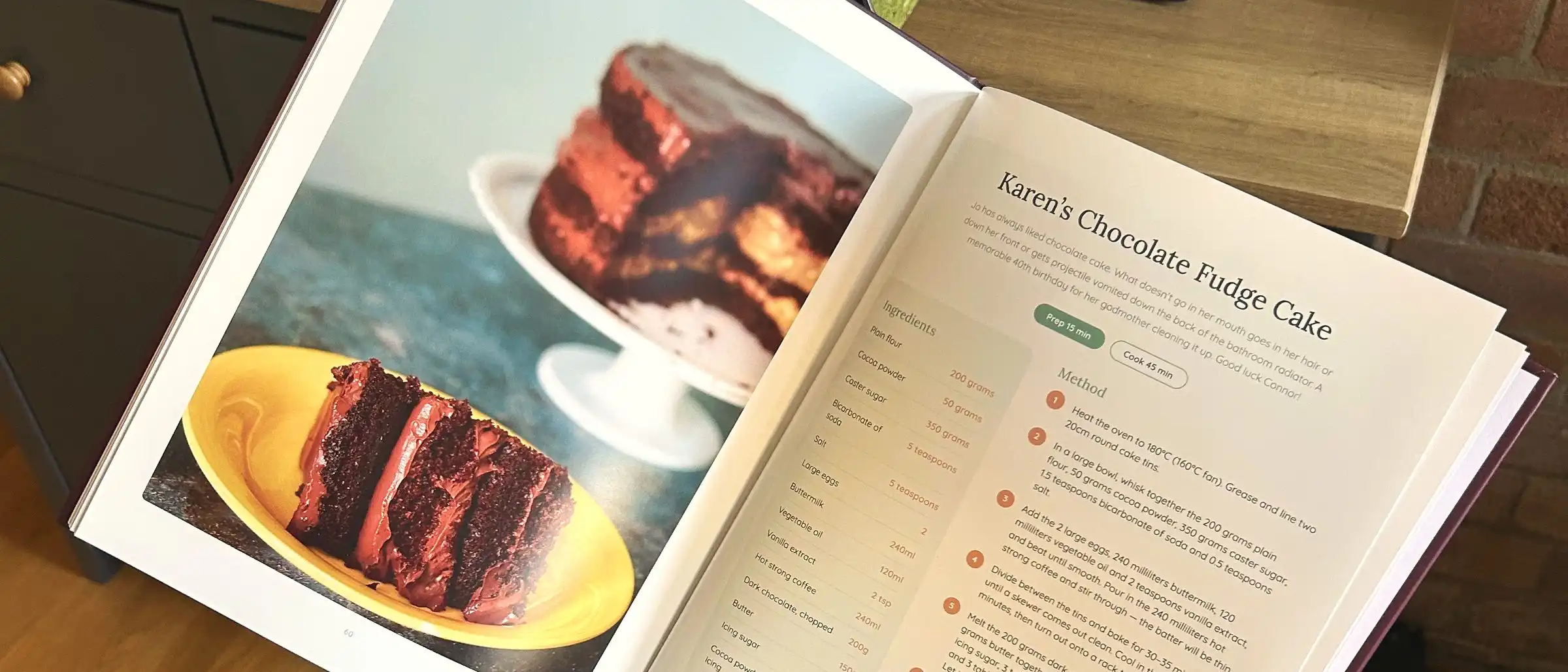 An open milestone-birthday cookbook showing a chocolate fudge cake recipe beside a full-page photo of the cake